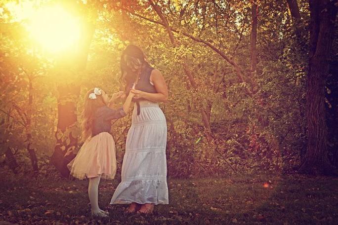 A young girl looks up at her mother in a wooded setting with the setting sun casting a golden light through the trees