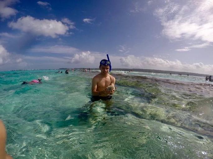 A young man wearing goggles and a snorkle on his forehead looks at a seacreature he has found in waist-deep waters.