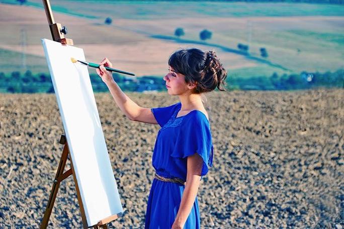 A woman in a blue dress paints standing up in a field in the countryside.